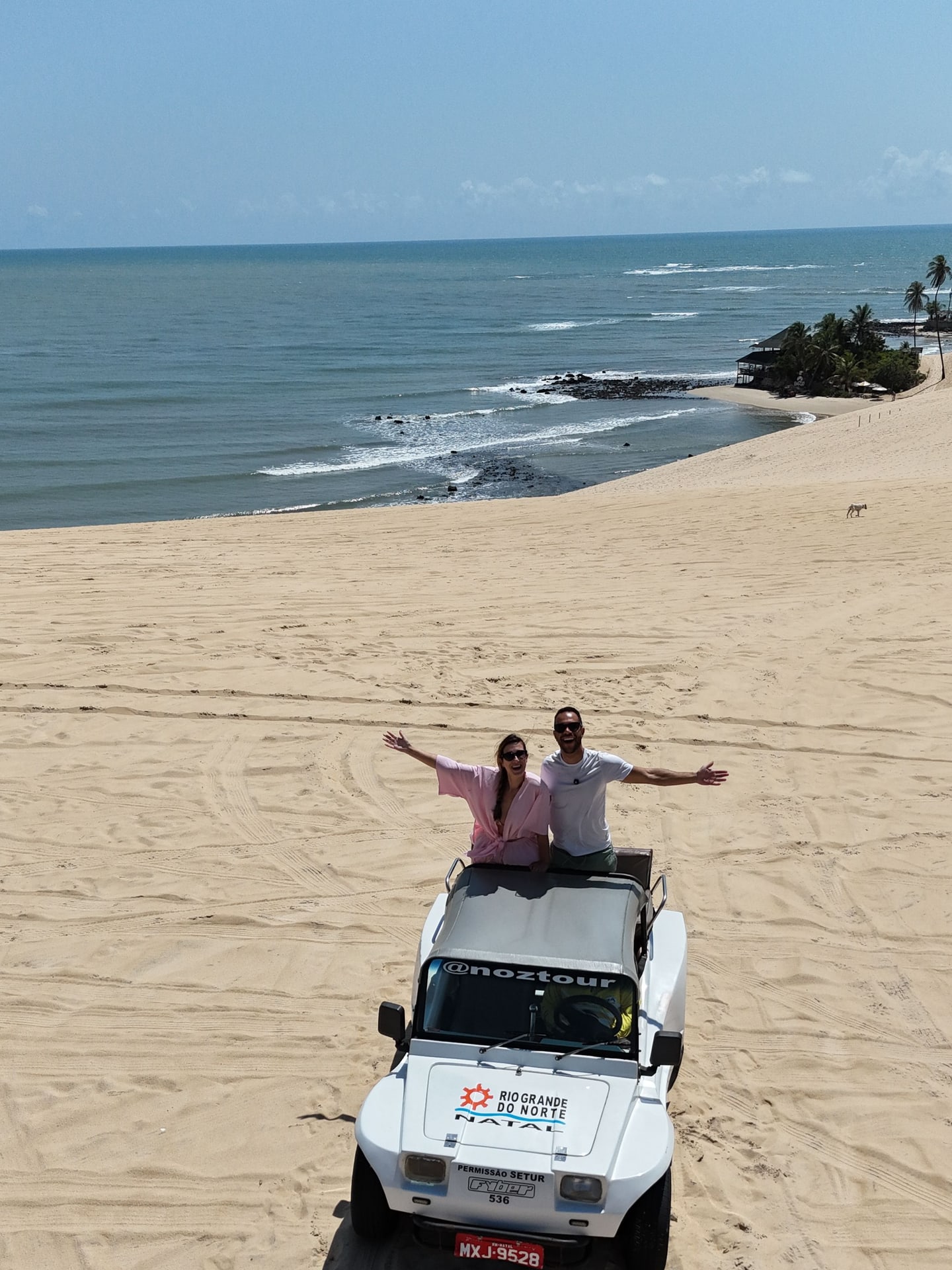Casal acenando em pé no buggy nas dunas do litoral norte de Natal, com o mar ao fundo