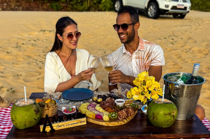 Casal brindando no piquenique nas dunas com água de coco e frutas