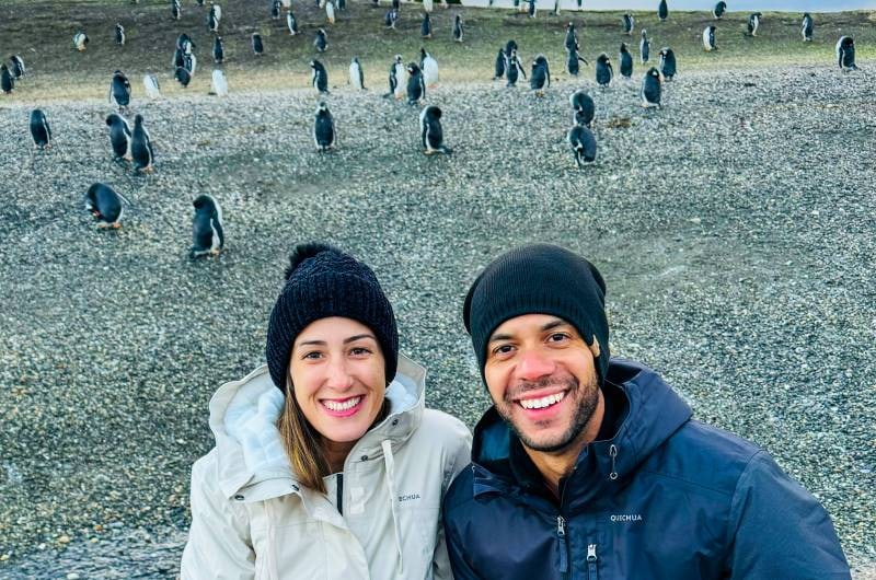 Casal no barco com a colônia de pinguins da Ilha Martillo ao fundo