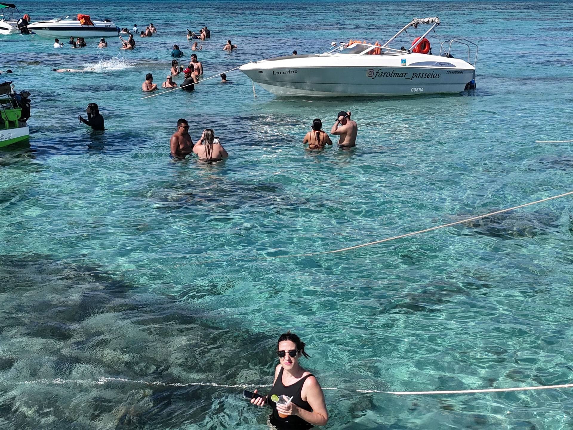 Pessoas na água rasa e transparente dos parrachos de Rio do Fogo, com lanchas ao fundo