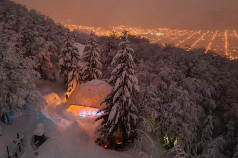 Refúgio iluminado na neve à noite no Cerro Otto, com as luzes de Bariloche ao fundo