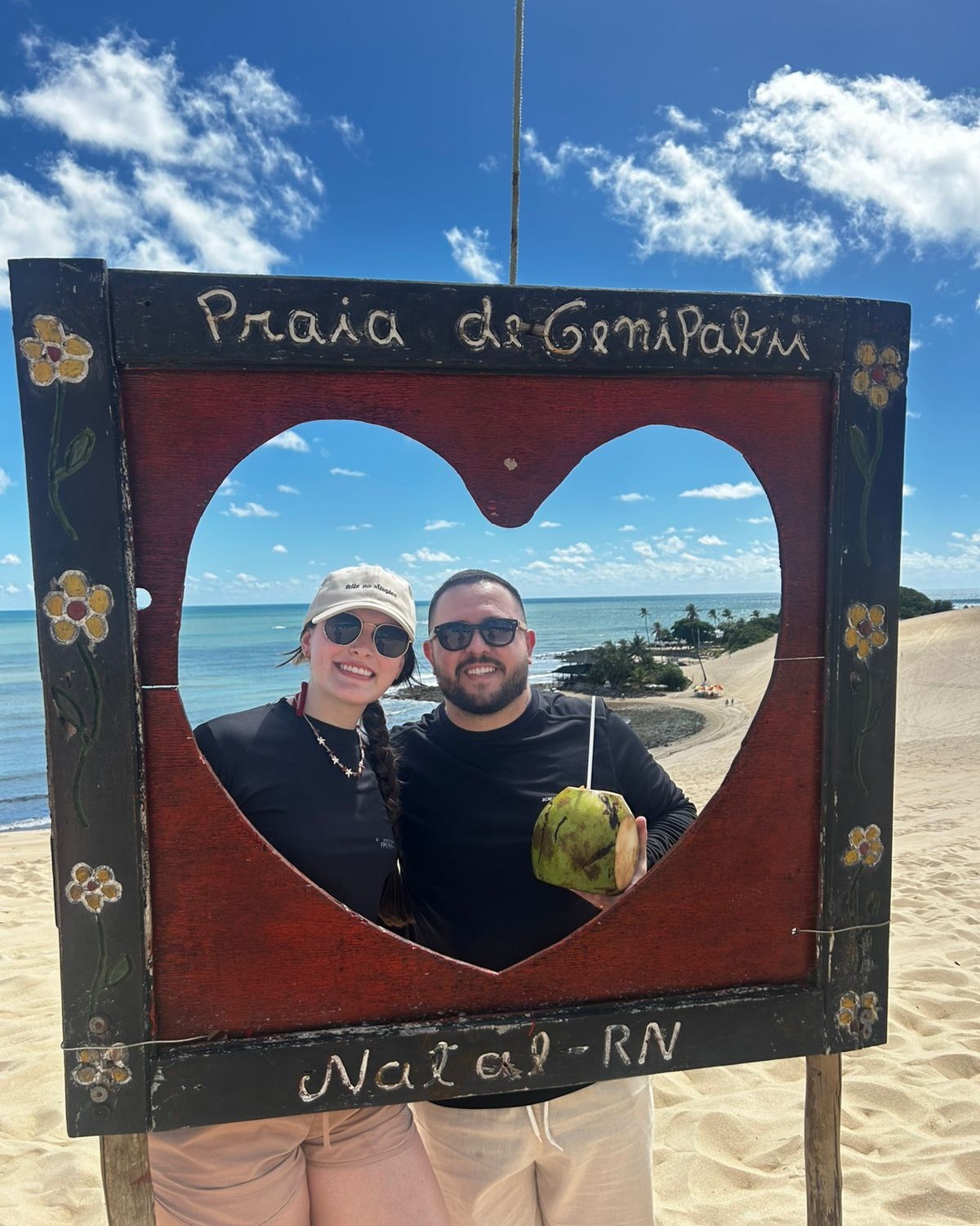 Casal sorrindo atrás da moldura de madeira em forma de coração da Praia de Genipabu, em Natal