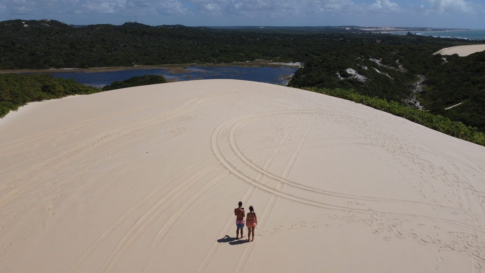 Casal no alto das Dunas de Genipabu, com marcas de buggy na areia e a lagoa e a mata ao fundo, visto de cima