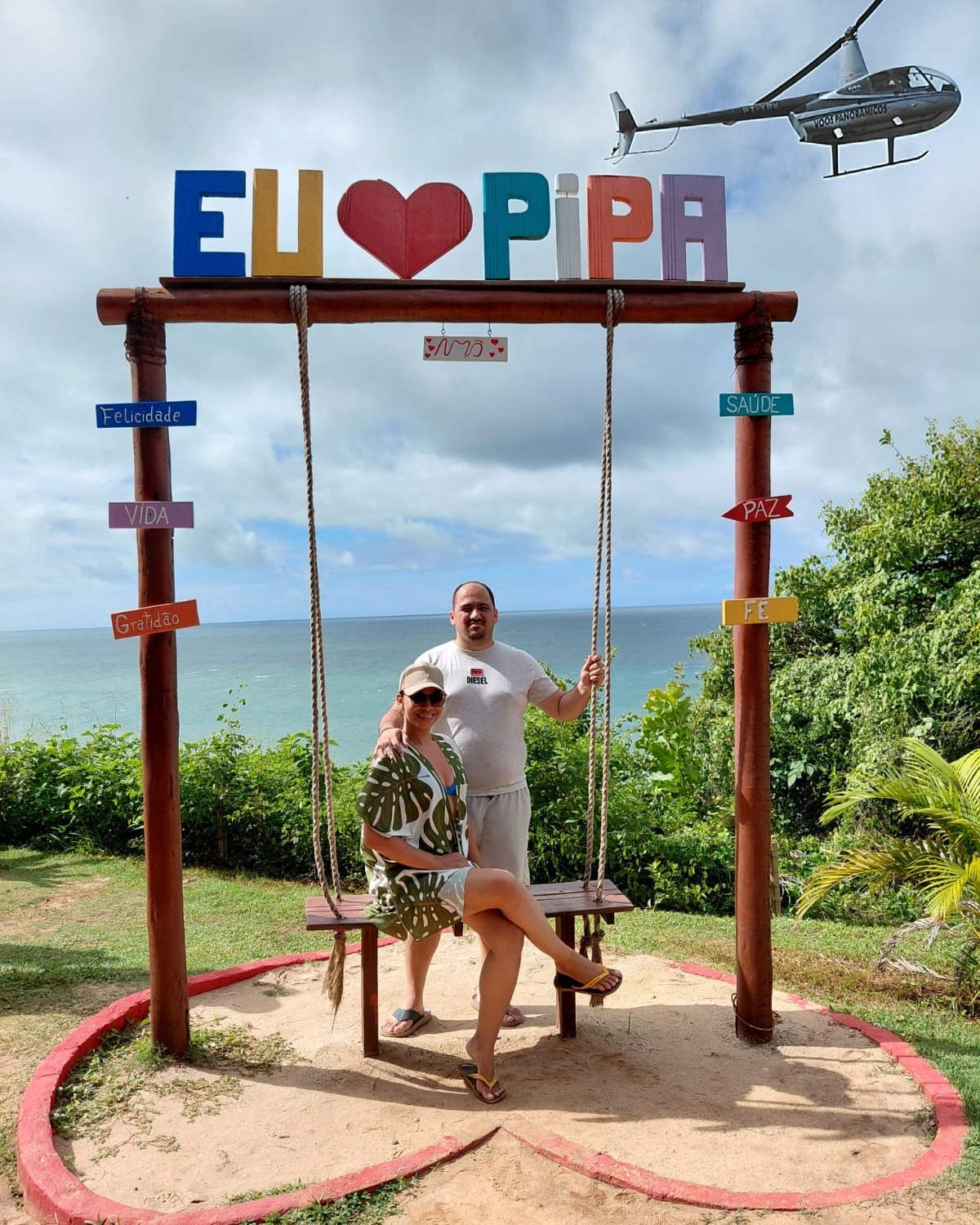 Casal no balanço com o letreiro Eu amo Pipa, num mirante com vista para o mar