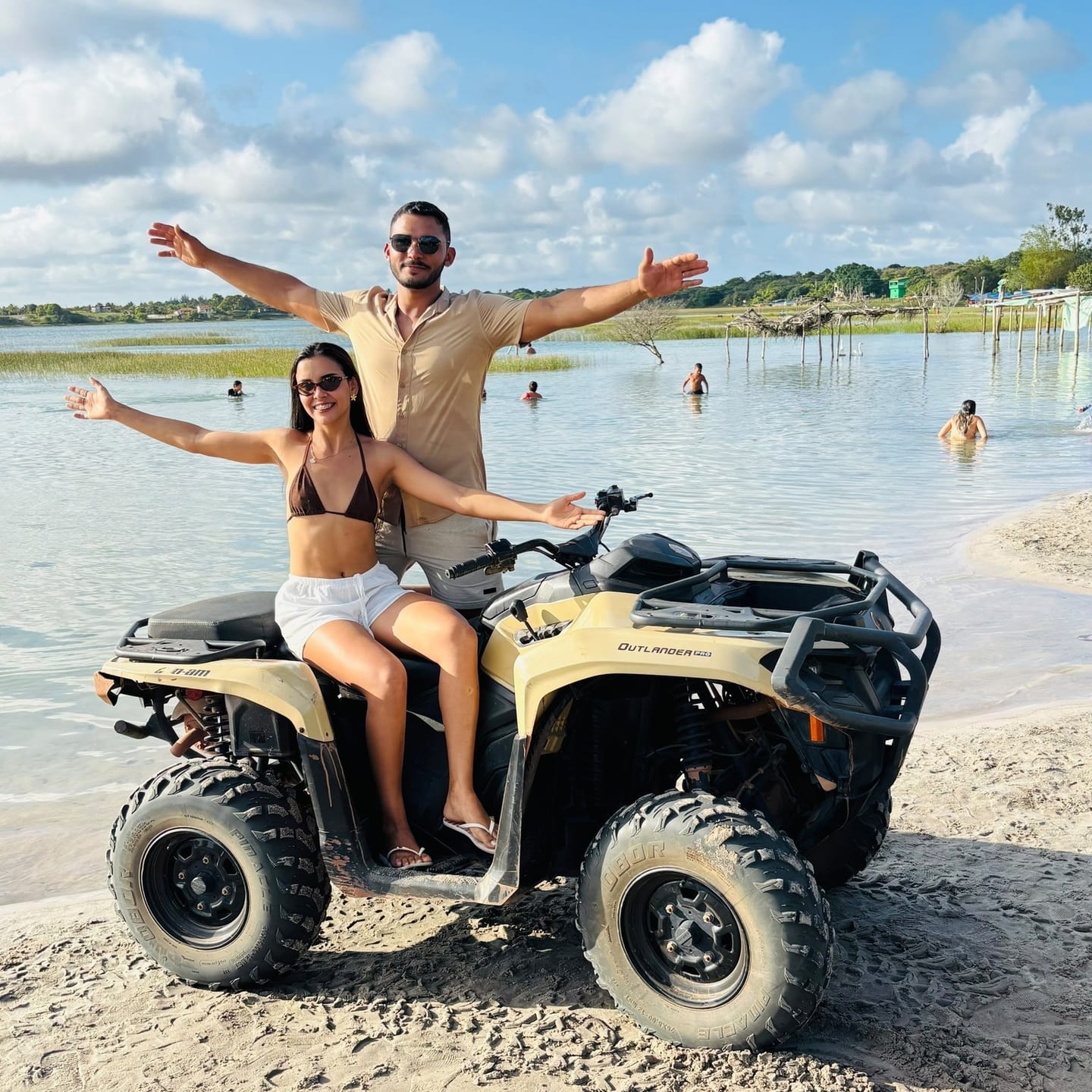 Casal de braços abertos em cima de um quadriciclo, na areia à beira de uma lagoa rasa