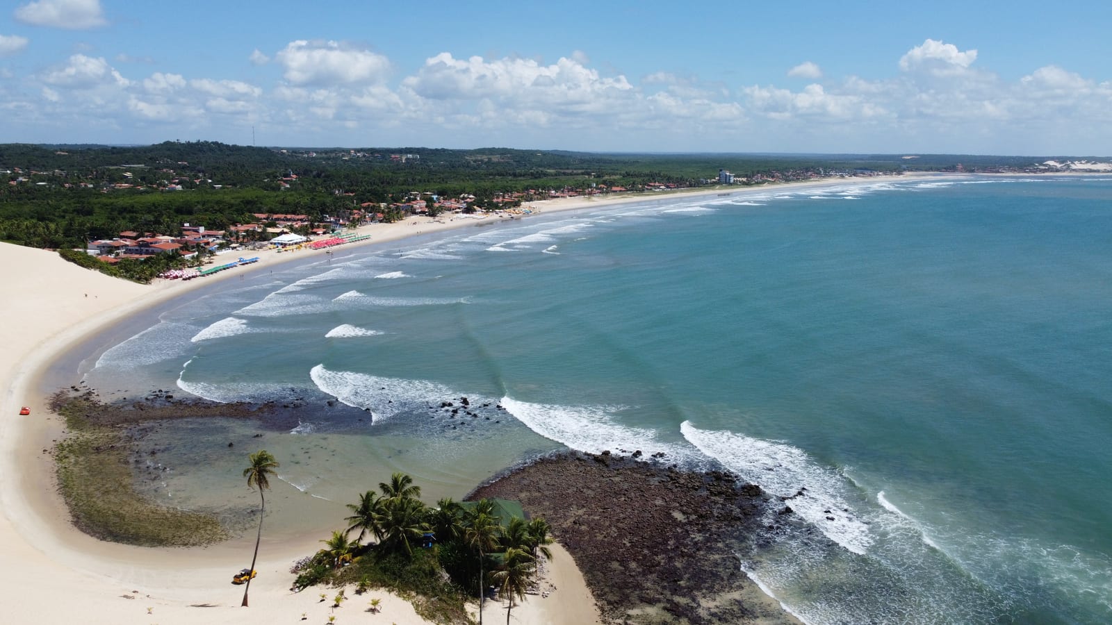 Duna de areia clara descendo até a praia de Genipabu, com coqueiros, recifes e o mar azul, vista de cima