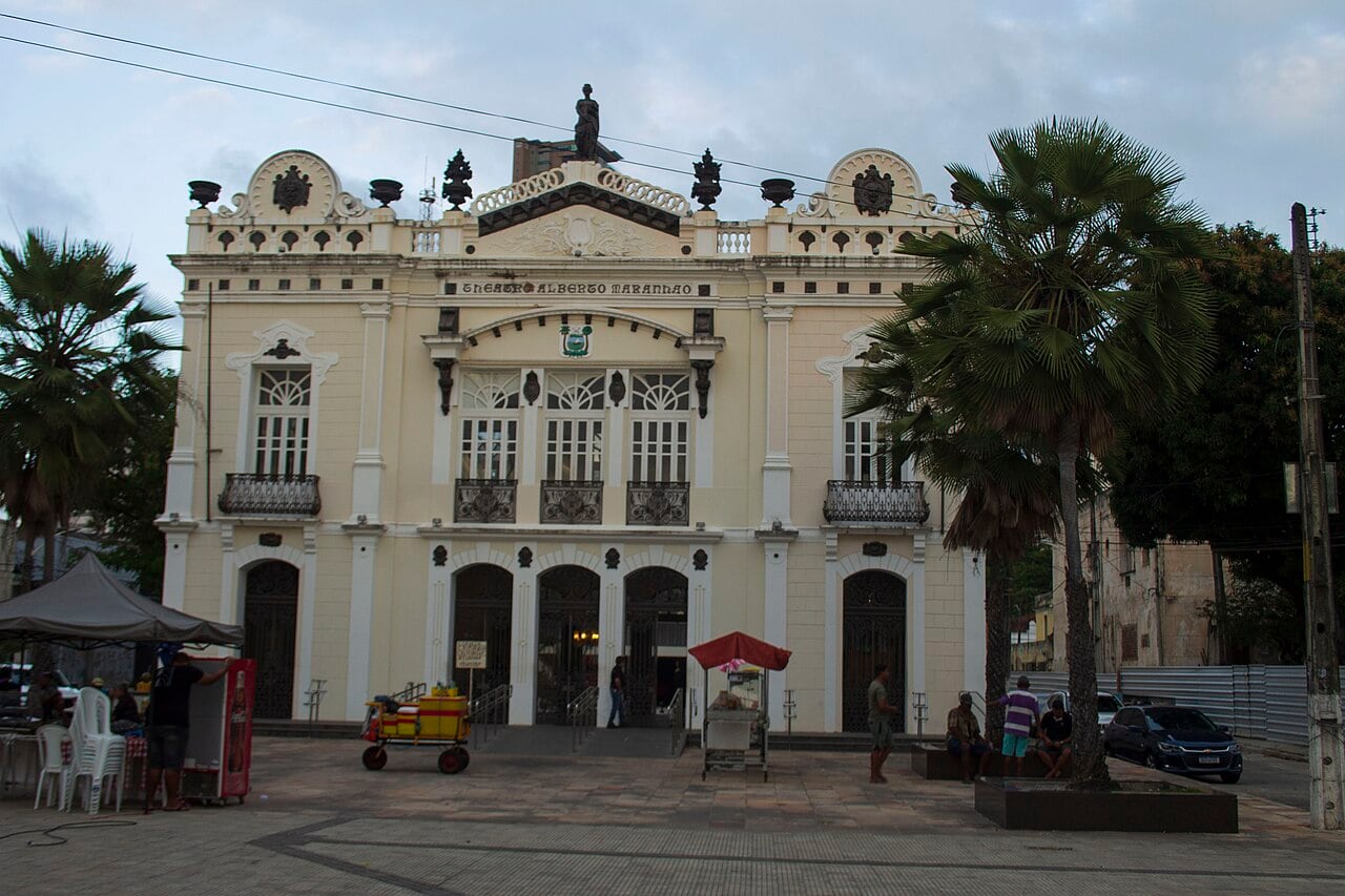 Fachada clara e ornamentada do Teatro Alberto Maranhão, com palmeiras e barracas de lanche na praça em frente, na Ribeira