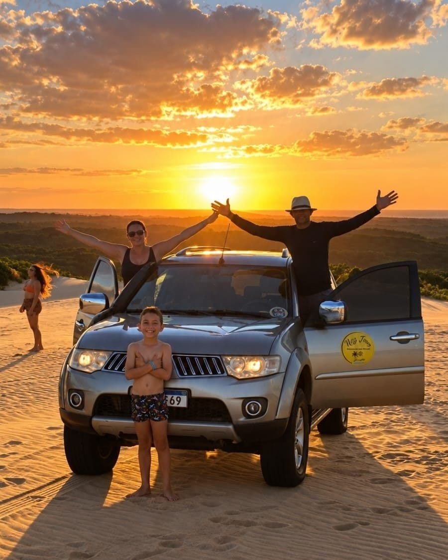 Família de braços abertos em volta da caminhonete 4x4 nas dunas, com o sol se pondo atrás