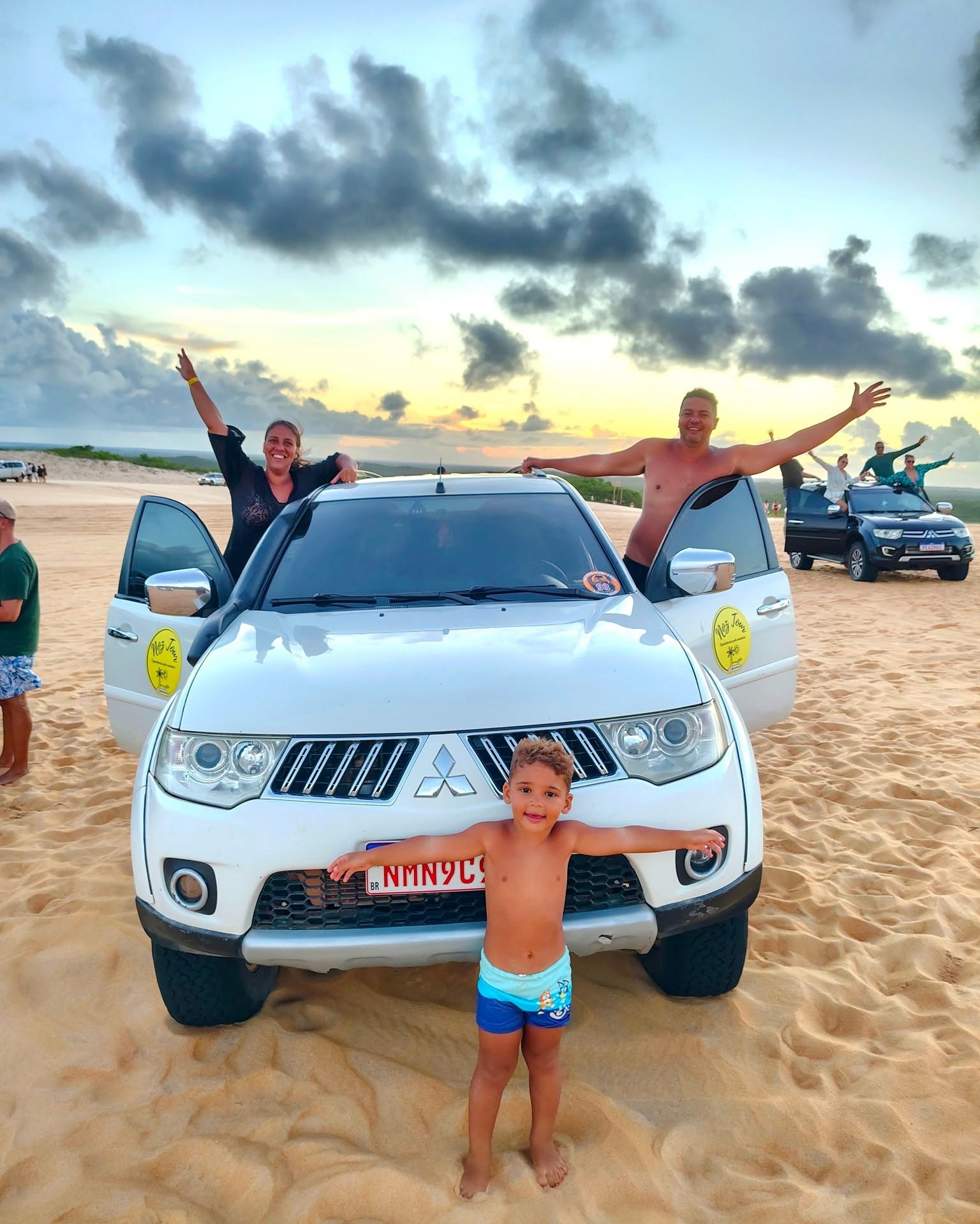 Família de braços abertos ao redor da caminhonete 4x4 nas dunas ao entardecer, em Natal