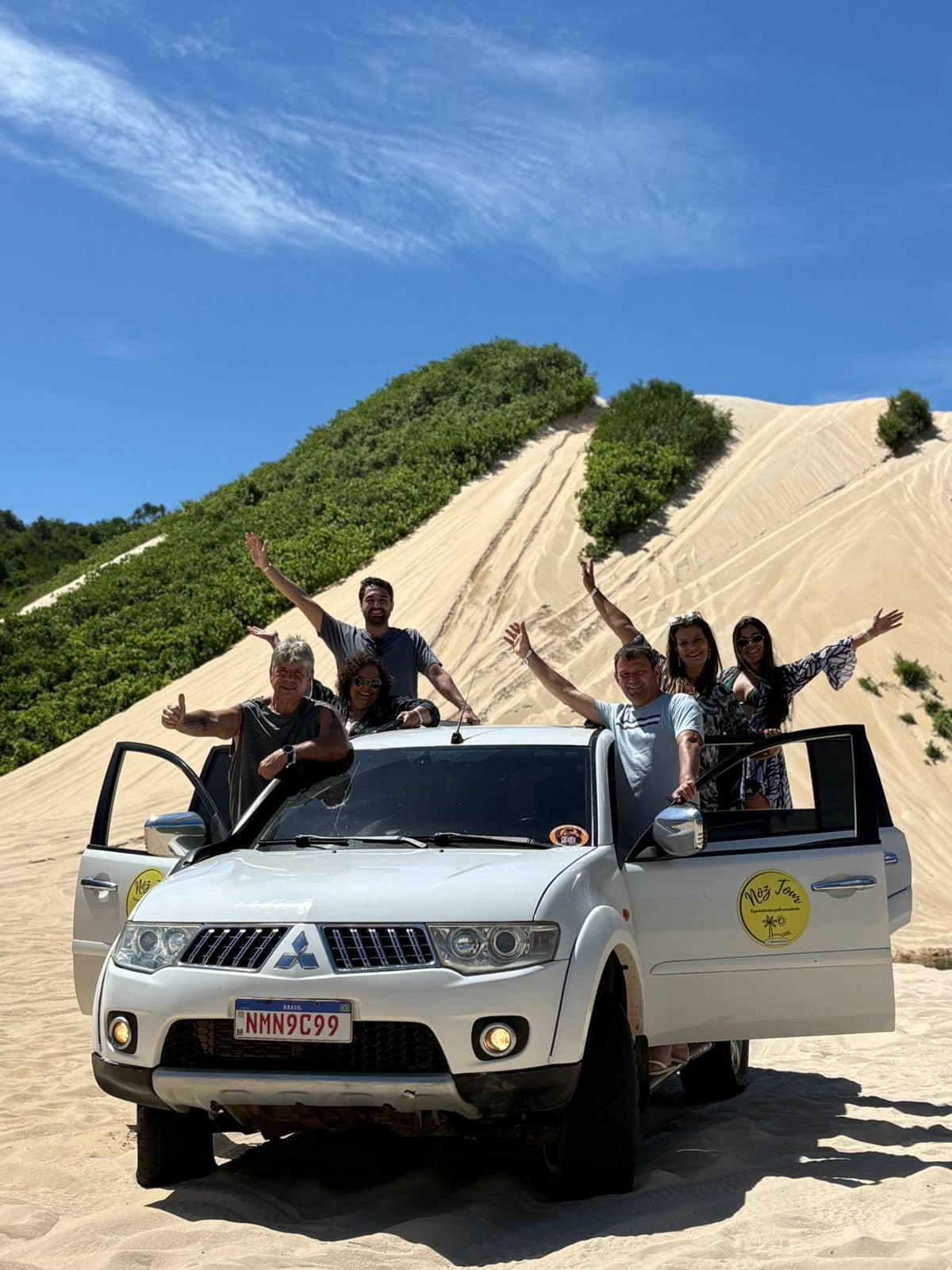 Grupo comemorando nas portas abertas de uma caminhonete 4x4 branca, parada no pé de uma duna, em Natal