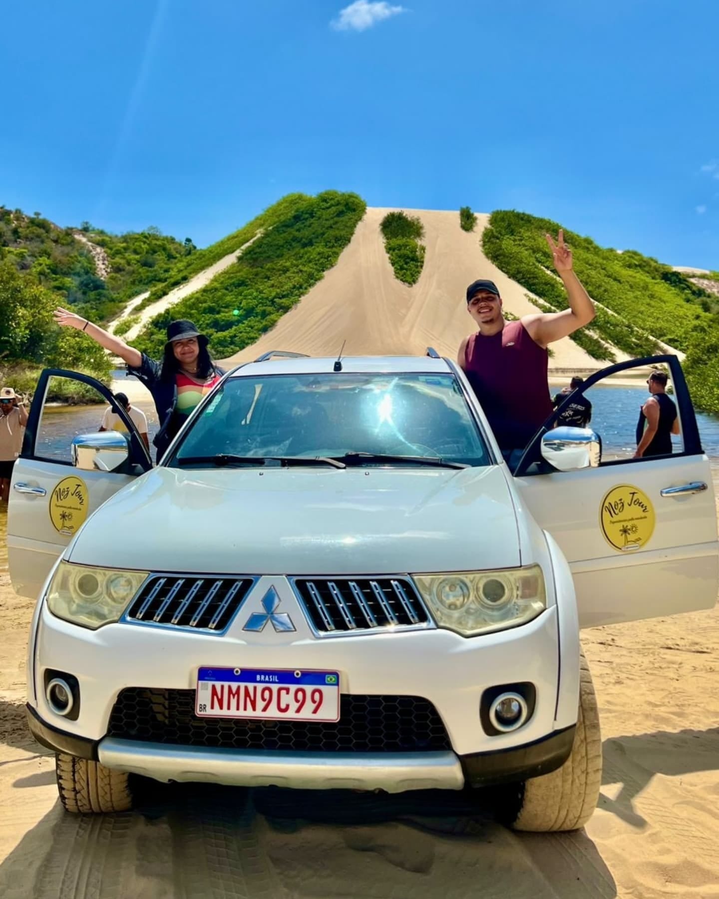 Grupo de viajantes acenando ao lado da caminhonete 4x4 parada na areia, entre dunas e vegetação