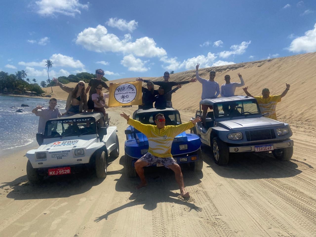 Grupo de viajantes comemorando em pé em três buggys estacionados na areia, perto do mar, em Natal