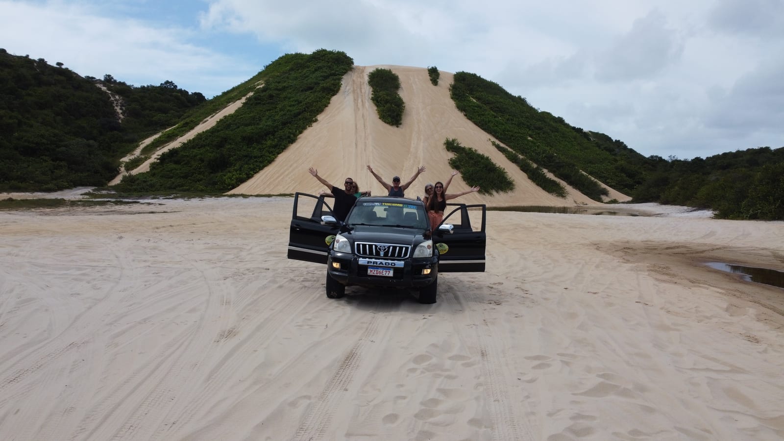 Grupo de braços abertos na caminhonete 4x4 de portas abertas, parada na areia ao pé de uma duna alta com mata dos dois lados