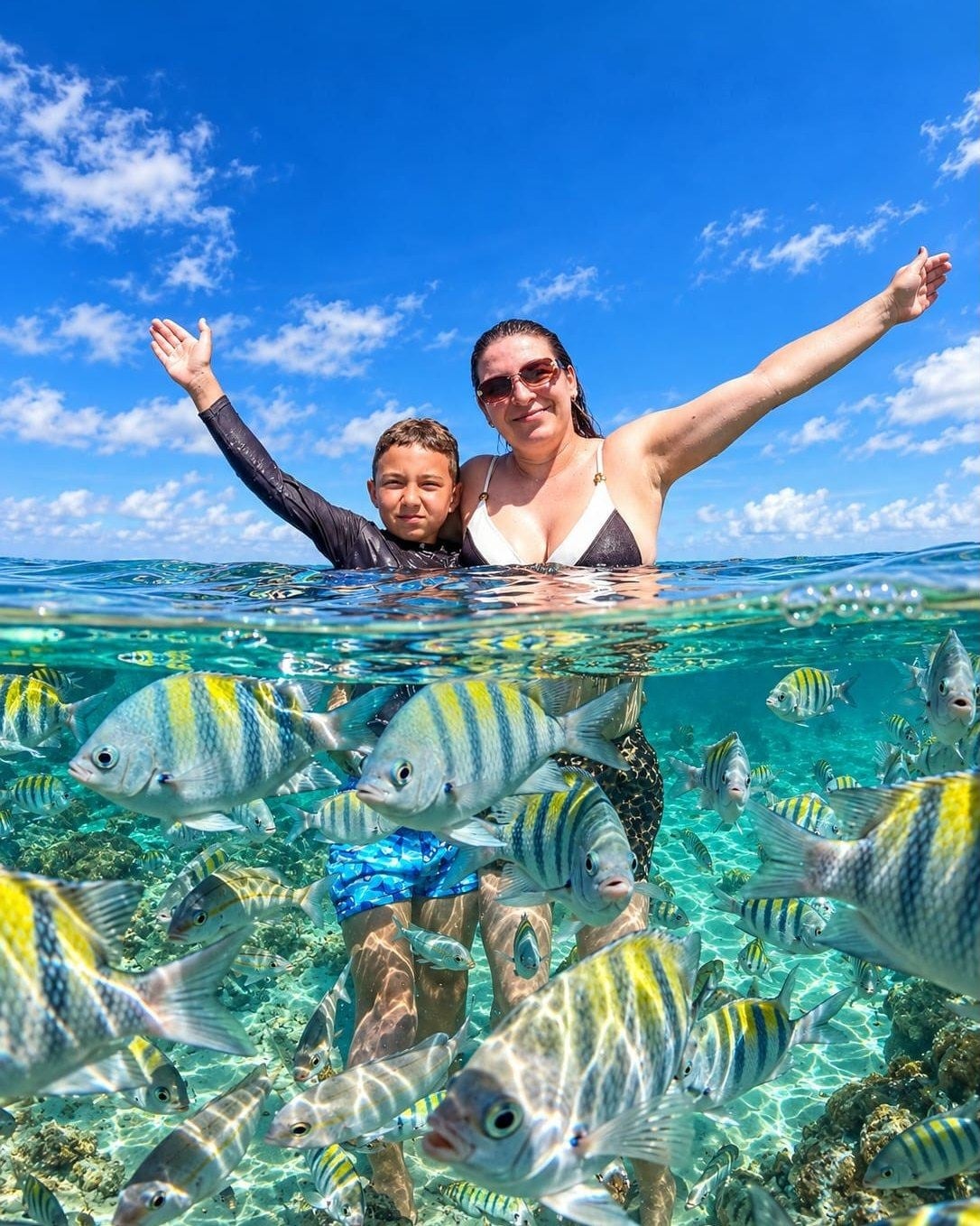 Mãe e filho sorrindo de braços abertos numa piscina natural rasa, com peixes listrados embaixo d'água