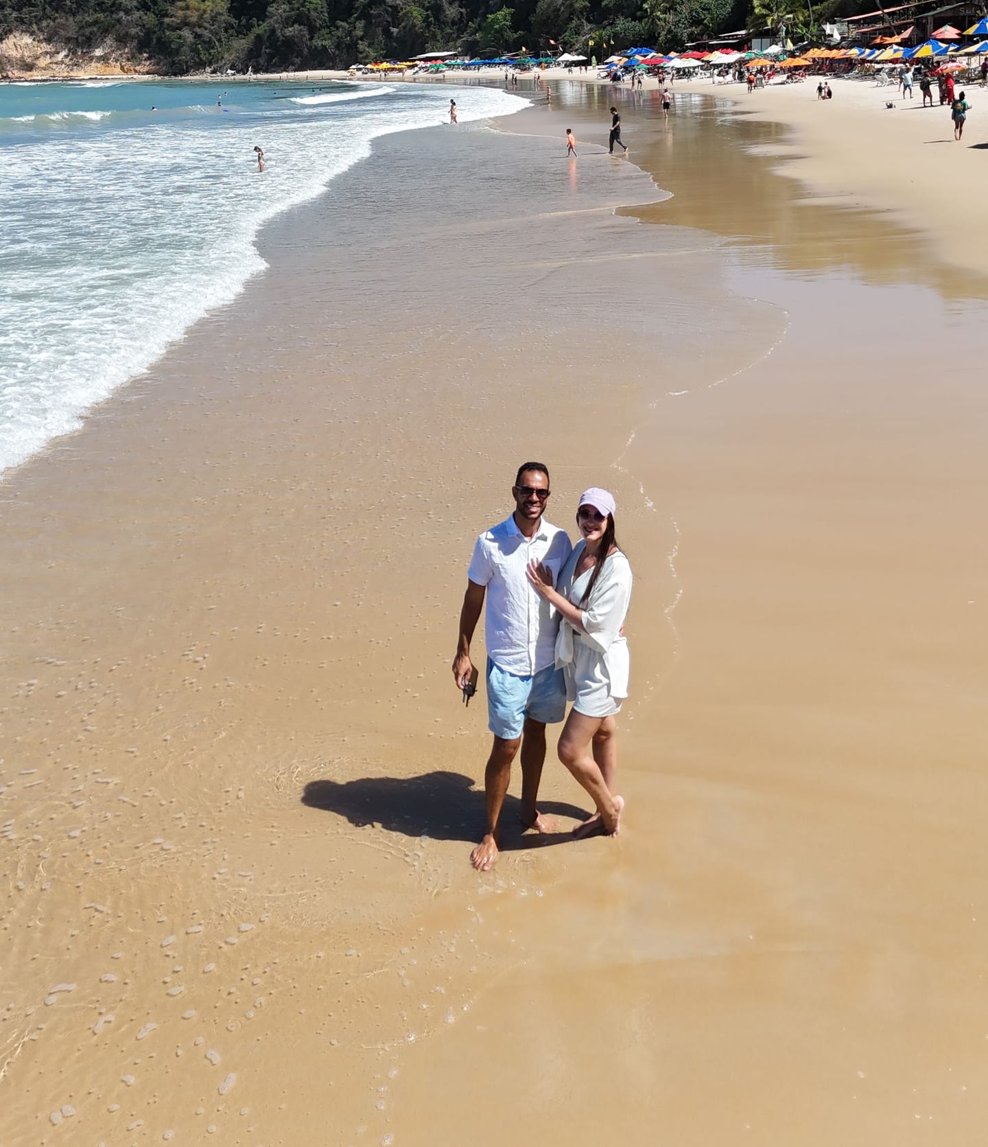 Monique e Robson abraçados na beira do mar numa praia de Pipa, com barracas coloridas e mata ao fundo