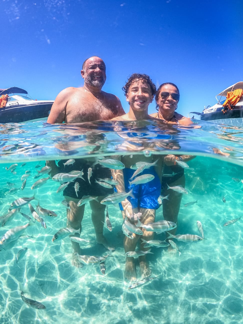 Três viajantes com a água pela cintura nos parrachos de Rio do Fogo, com peixes e lanchas ao redor