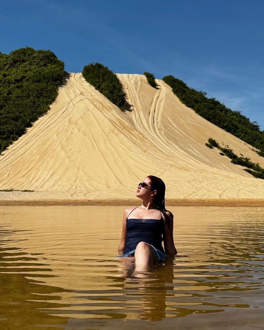 Viajante sentada na água rasa de uma lagoa, com uma duna alta de areia clara e vegetação atrás