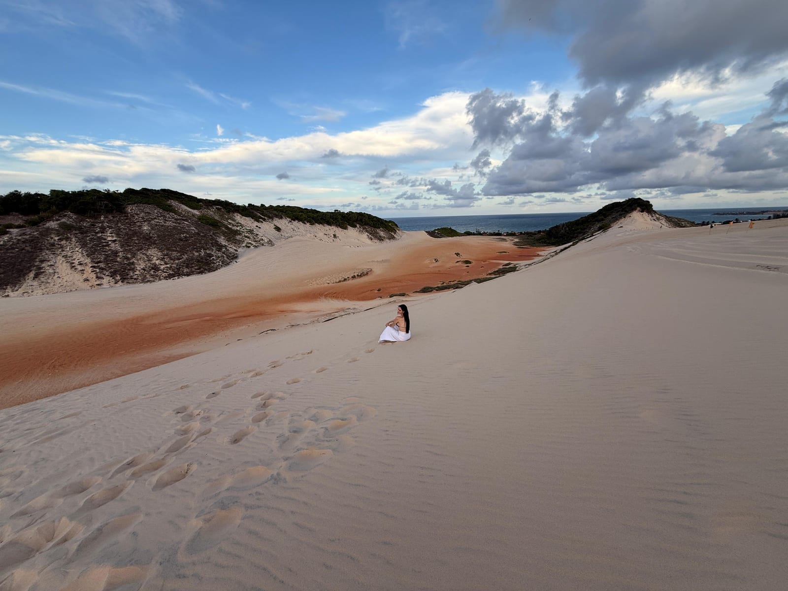 Viajante de vestido branco sentada na duna, com faixas de areia alaranjada, mata e o mar ao fundo