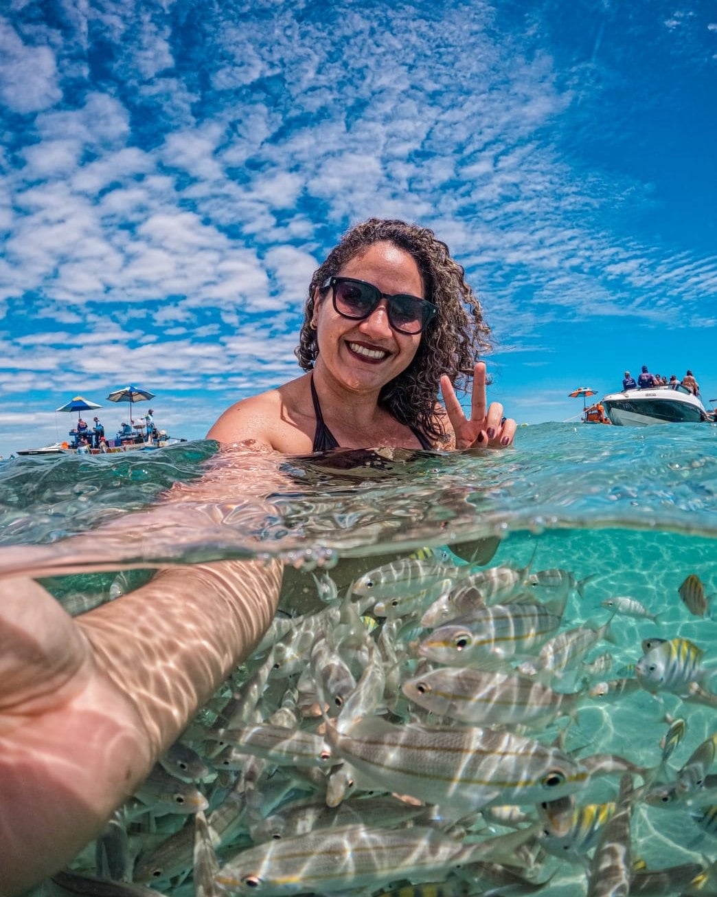 Viajante sorrindo na água transparente dos parrachos de Rio do Fogo, com peixes perto da câmera e lanchas ao fundo