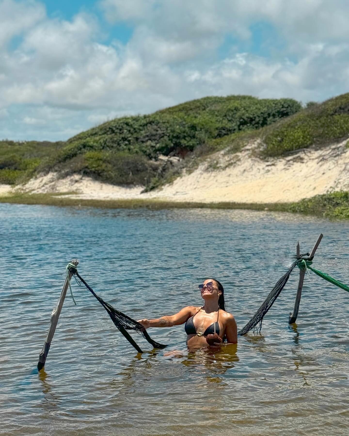 Viajante deitada numa redinha armada dentro da água, com duna e vegetação ao fundo