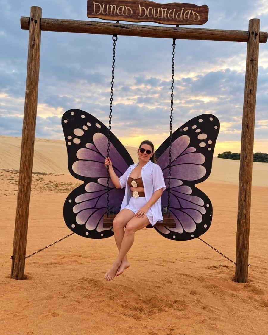 Viajante sentada num balanço com asas de borboleta na placa das Dunas Douradas, com a areia alaranjada ao fundo