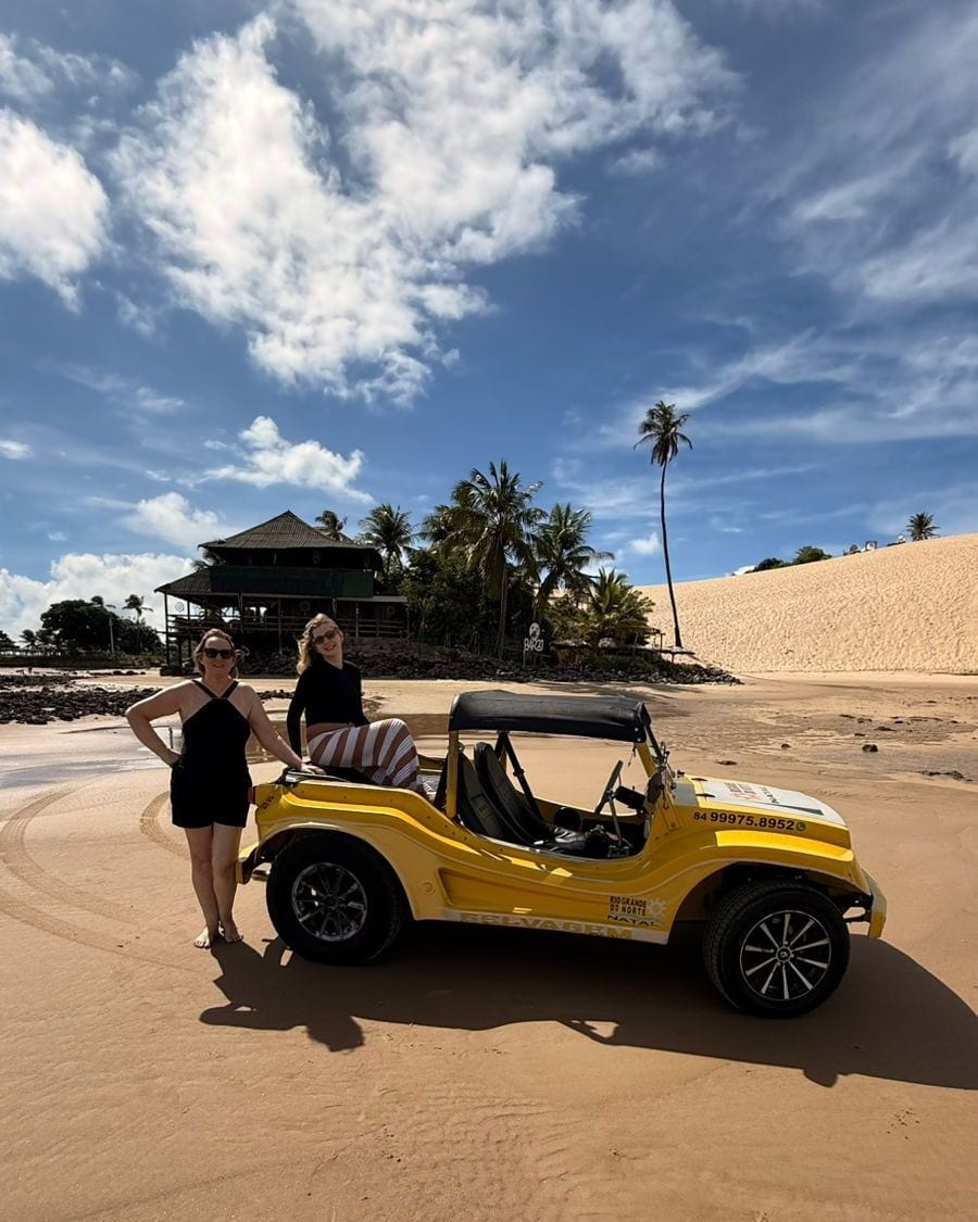 Duas viajantes ao lado de um buggy amarelo na areia, com dunas, coqueiros e céu azul ao fundo, em Natal