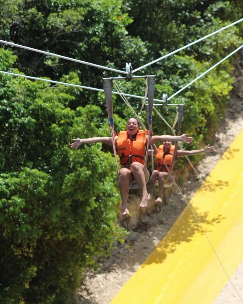 Dois viajantes de colete laranja descendo de tirolesa entre a mata, ao lado de uma rampa amarela