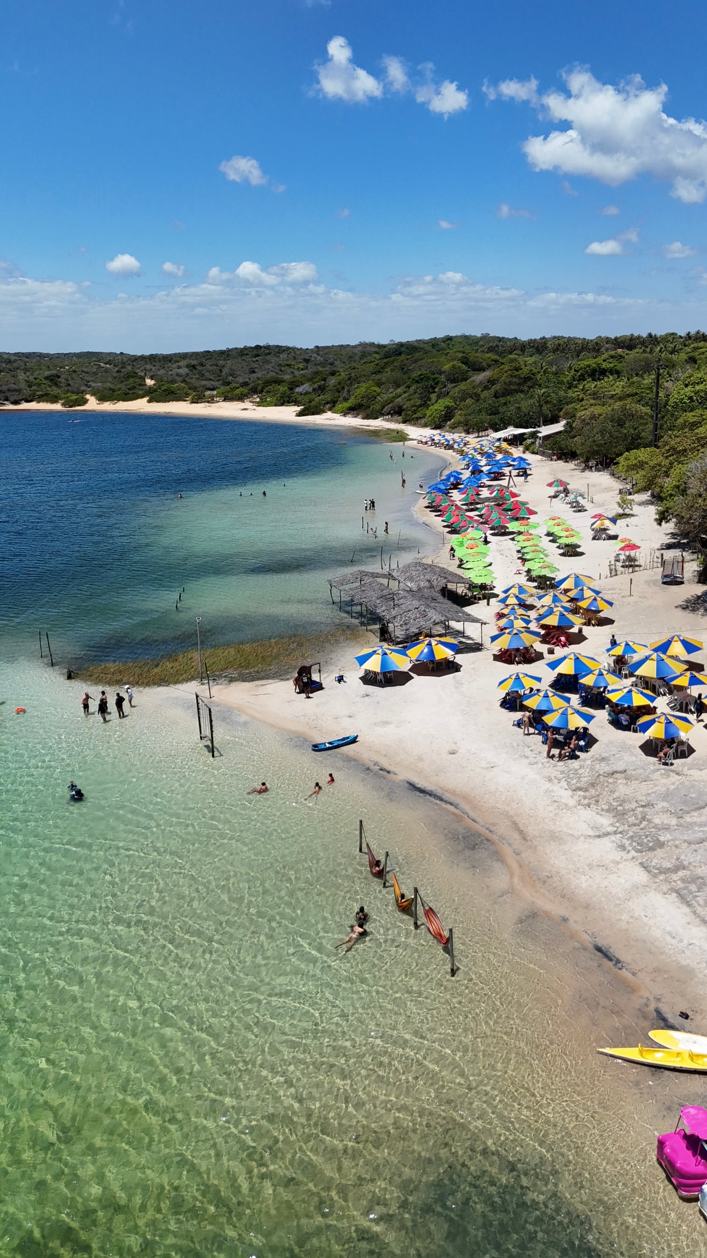 Vista aérea de uma lagoa de água verde clara com guarda-sóis coloridos e banhistas na margem, entre a mata