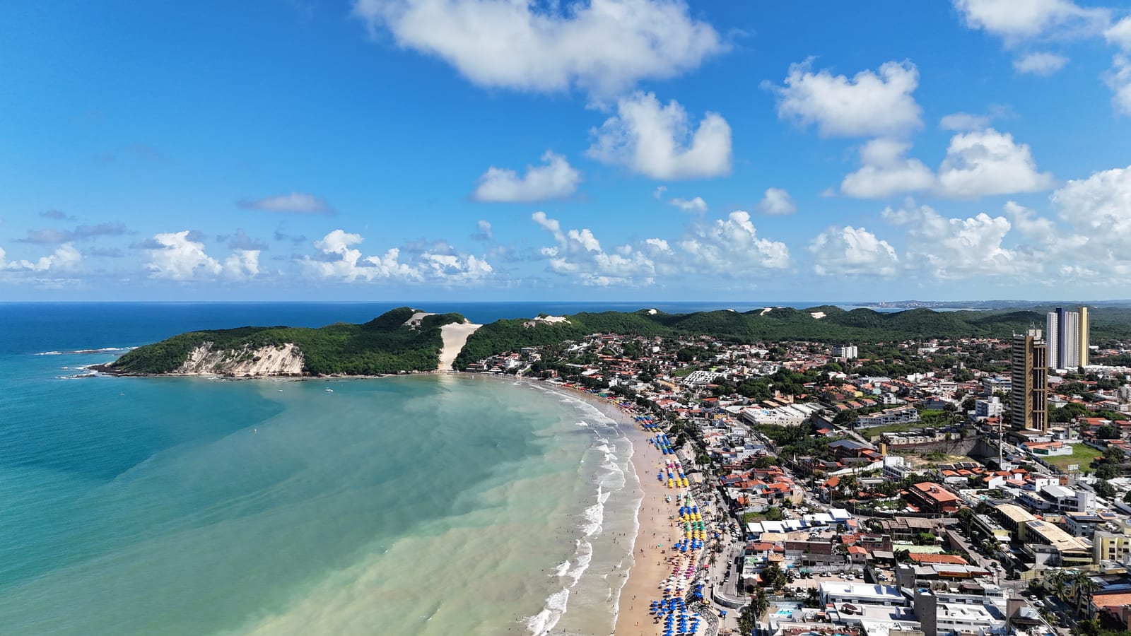 Vista aérea da Praia de Ponta Negra com guarda-sóis coloridos na areia e o Morro do Careca no fim da orla