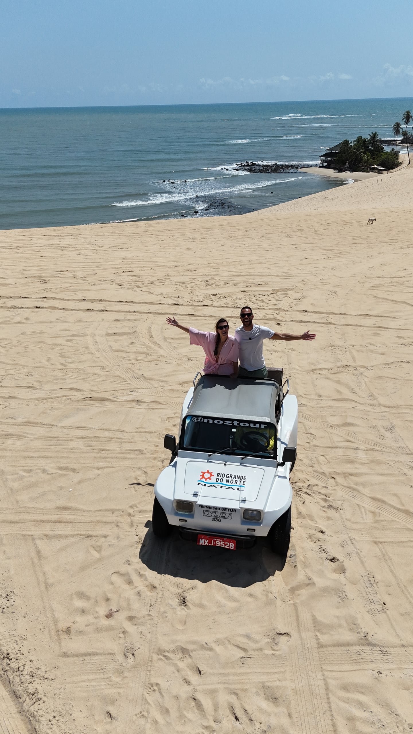 Vista aérea de um buggy branco com viajantes de braços abertos no alto de uma duna, com o mar ao fundo, em Natal